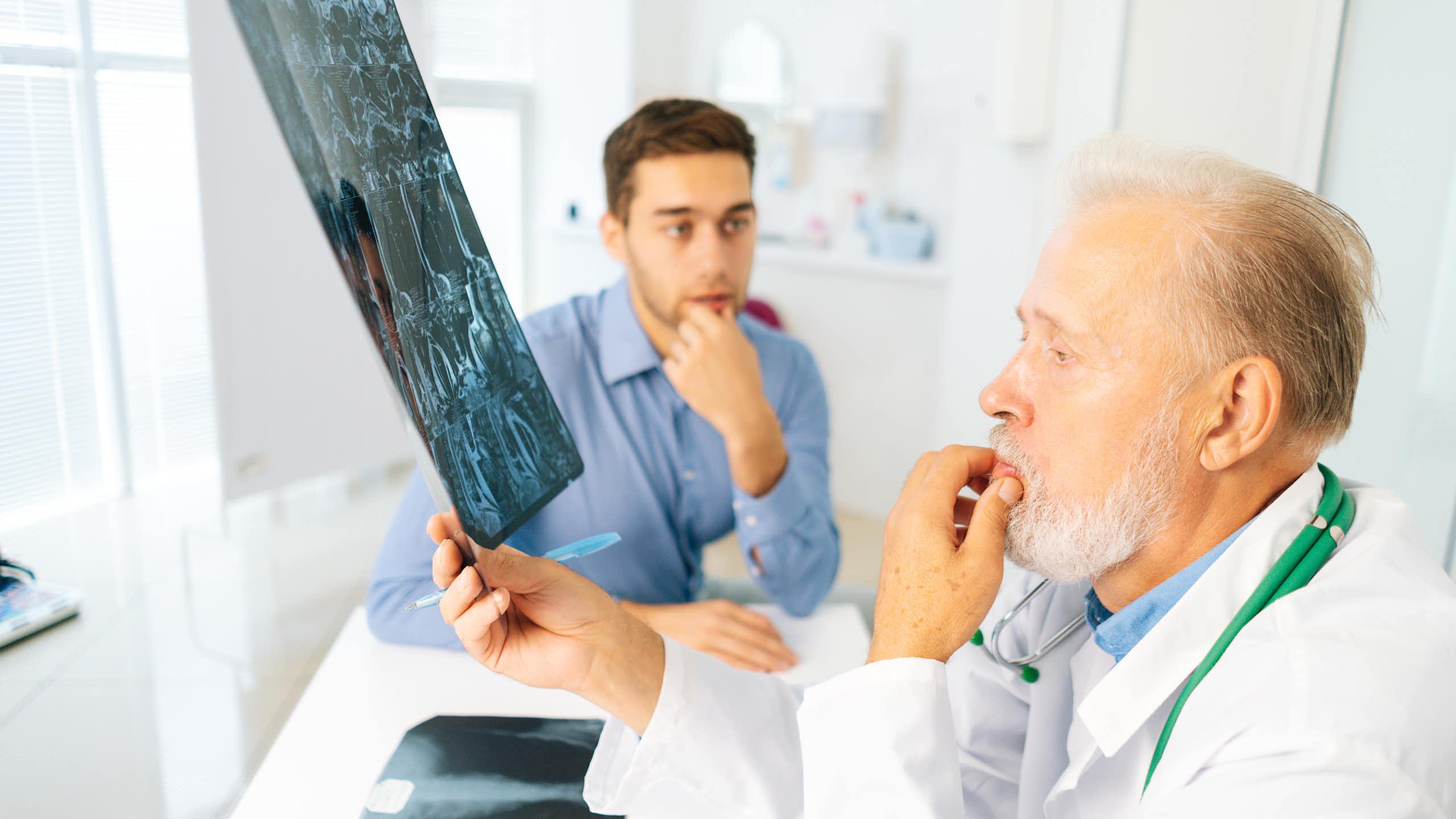 Un médico mira una radiografía de un joven (sentado al fondo con cara de preocupación).