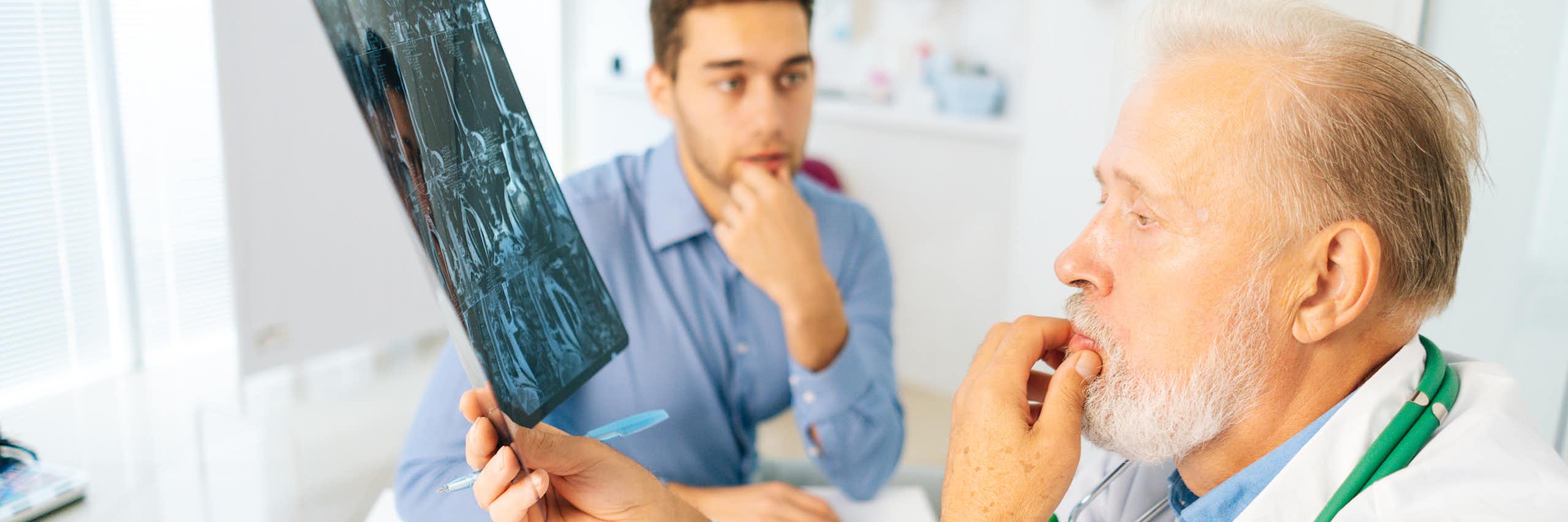 Un médico mira una radiografía de un joven (sentado al fondo con cara de preocupación).