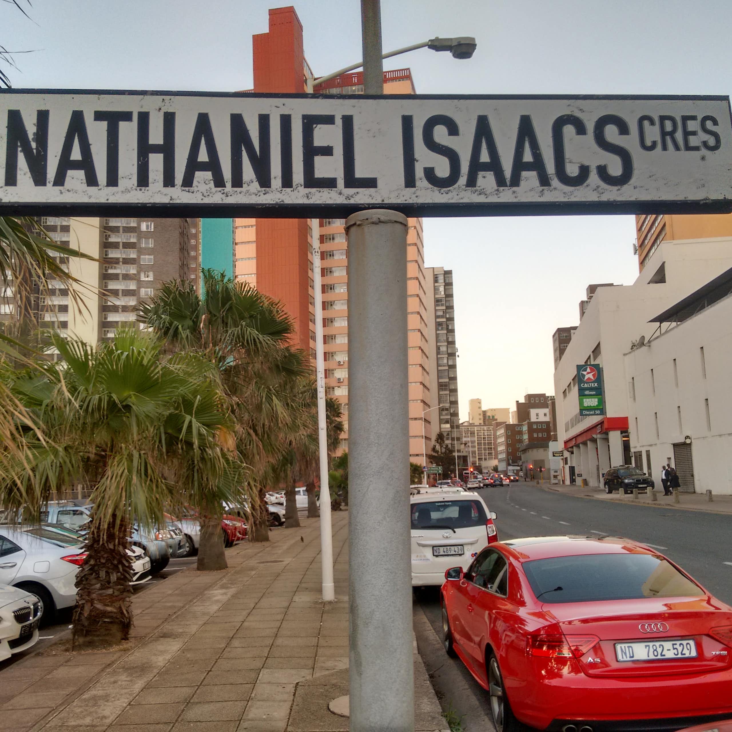 A sign saying 'Nathaniel Isaacs Cres' stands on a road with cars and high-rises in the background.
