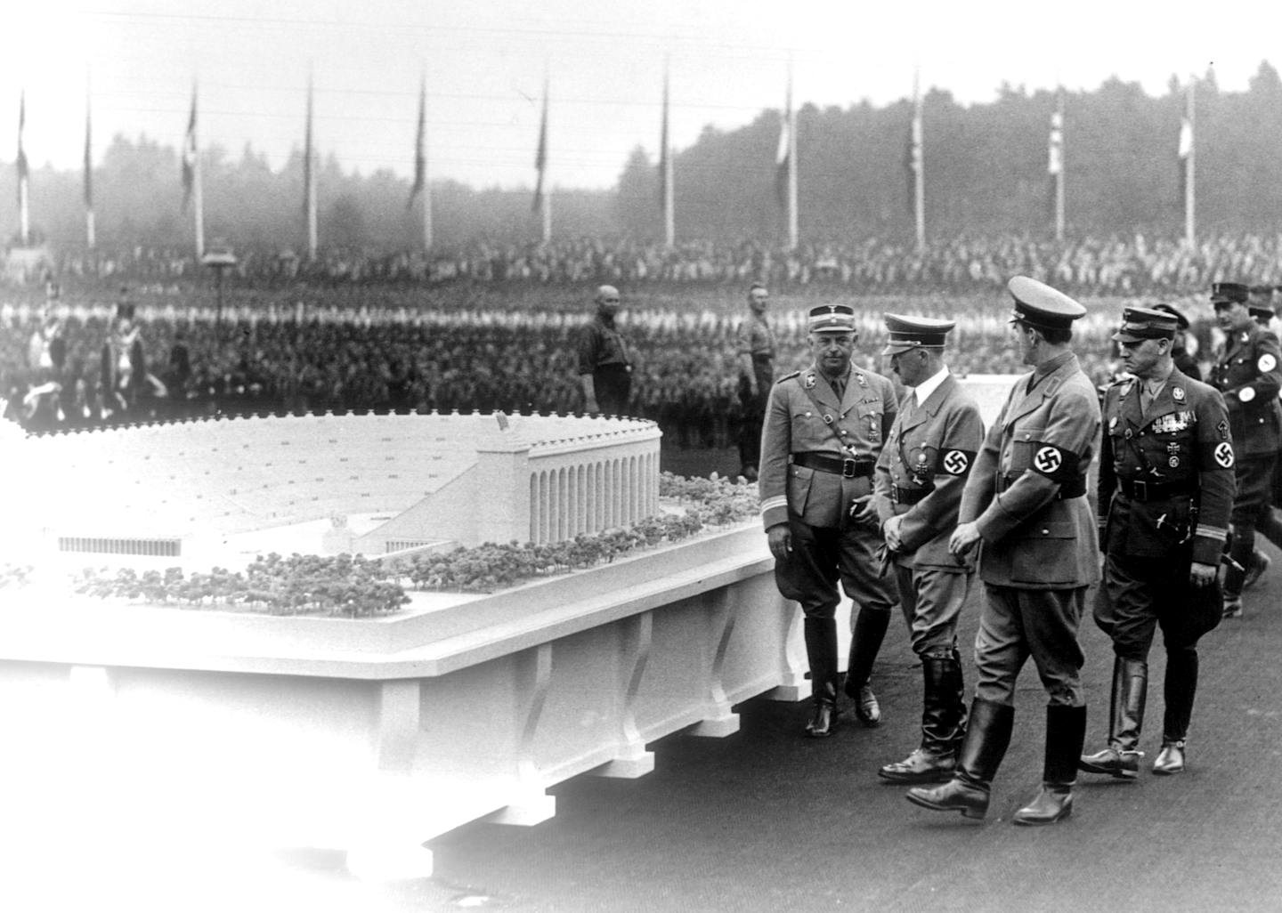 Four men in military uniforms with swastika arm bands inspect a large model of a stadium.