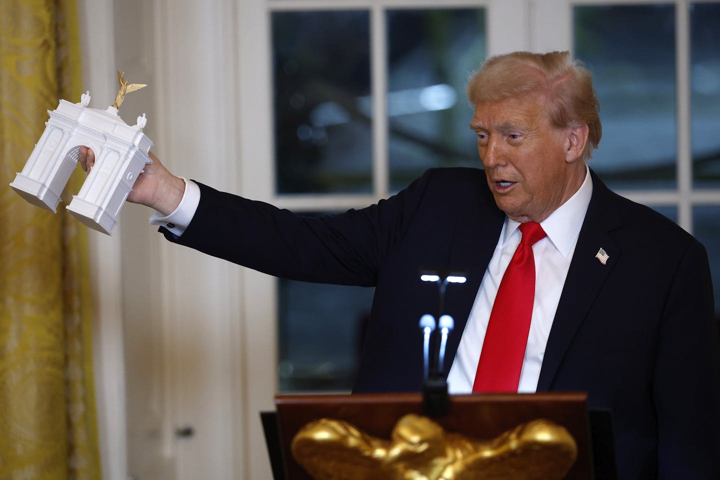 A man in a suit at a podium holds out a model of a white arch topped with a gilded statue.