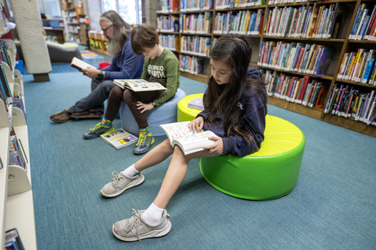 Two school-age children and one adult sit and read books, in front of a large shelf of books.