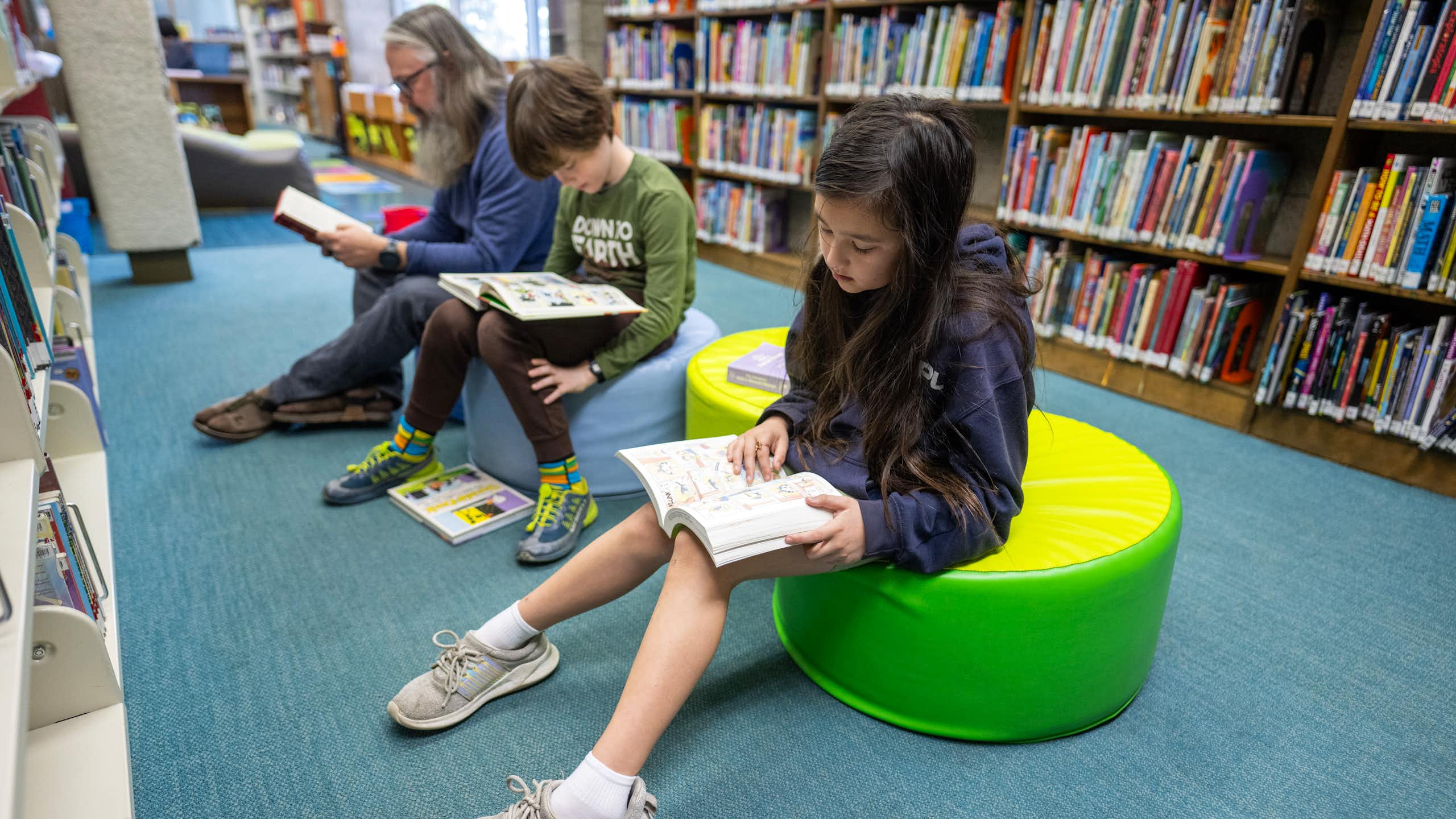 Two school-age children and one adult sit and read books, in front of a large shelf of books.