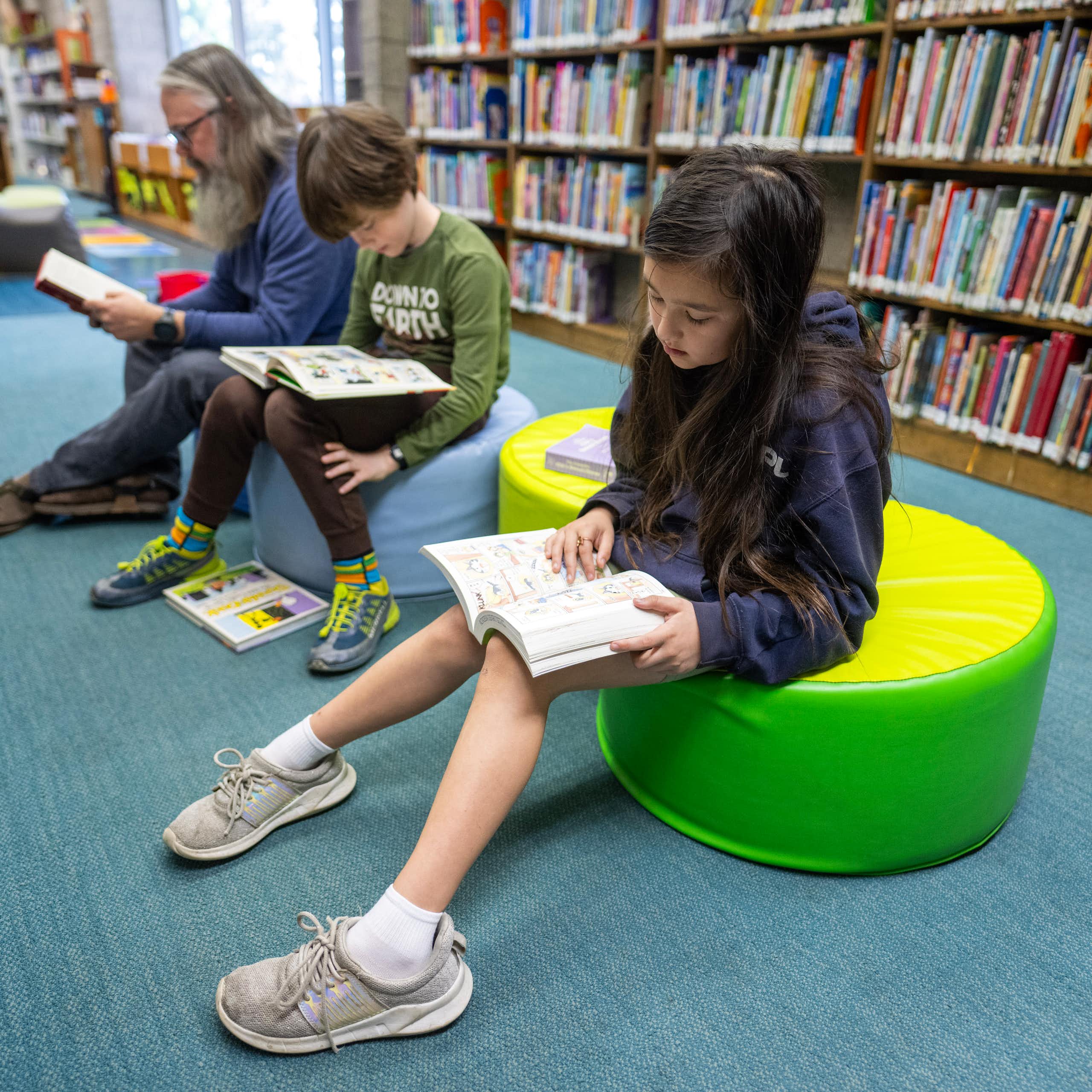 Two school-age children and one adult sit and read books, in front of a large shelf of books.