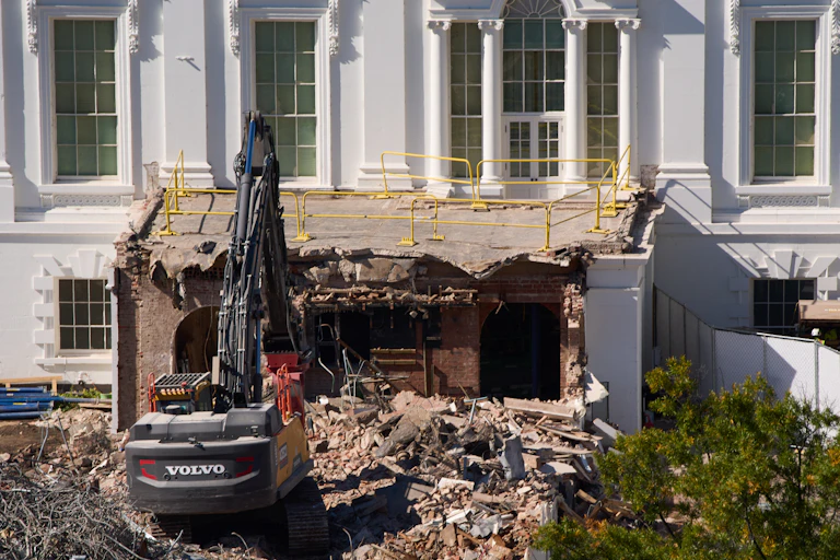 A construction machine pulls apart a section of a white building with columns above it.