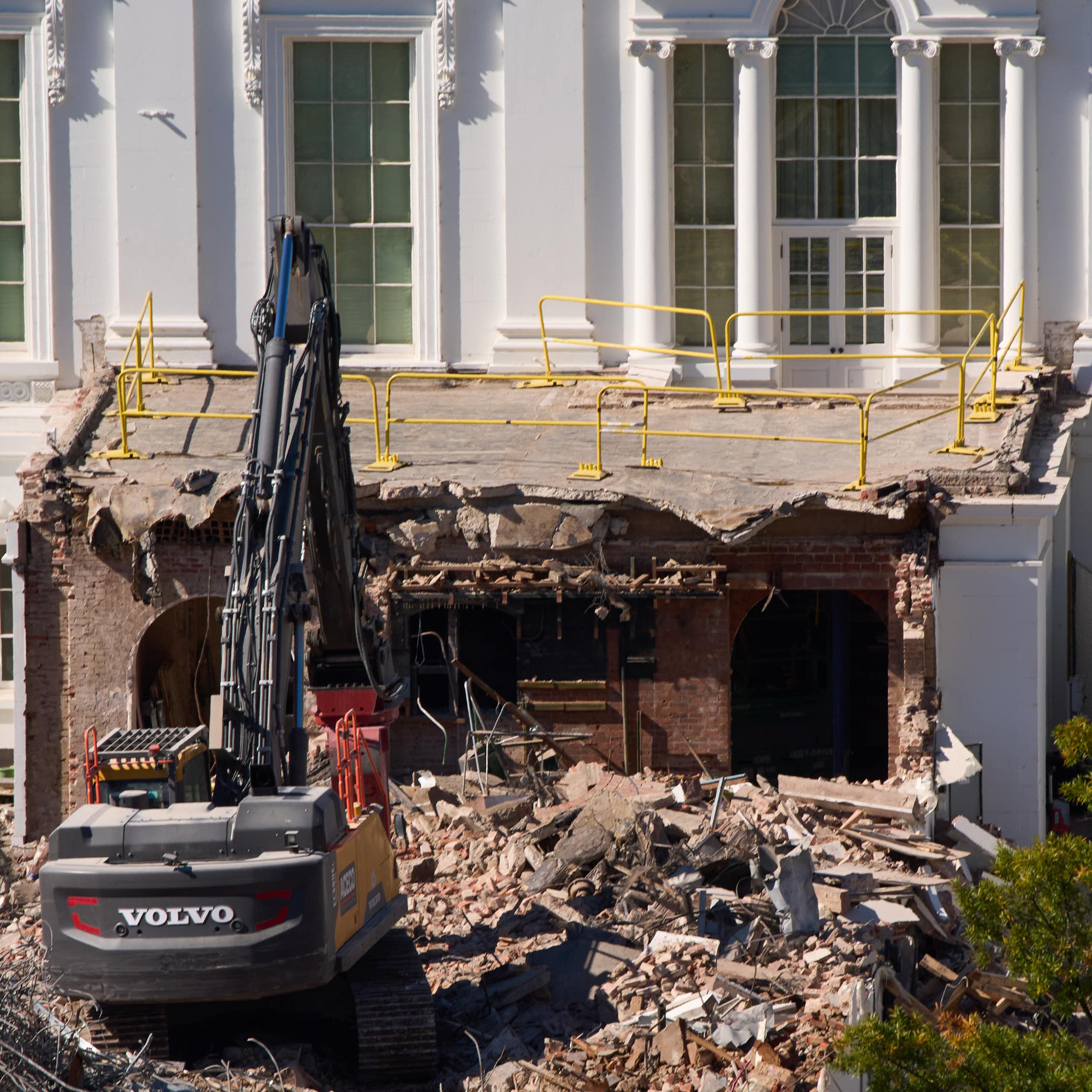 A construction machine pulls apart a section of a white building with columns above it.