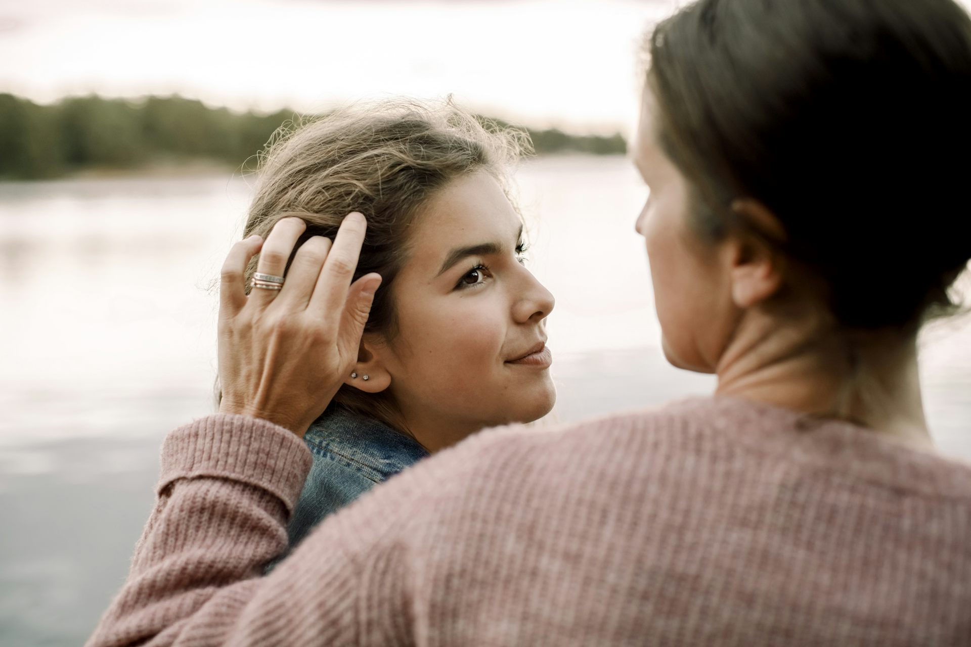 Woman tucks hair behind her daughter's ear as they stand next to a river.