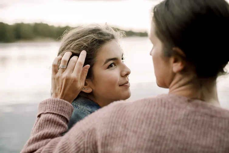 Woman tucks hair behind her daughter's ear as they stand next to a river.