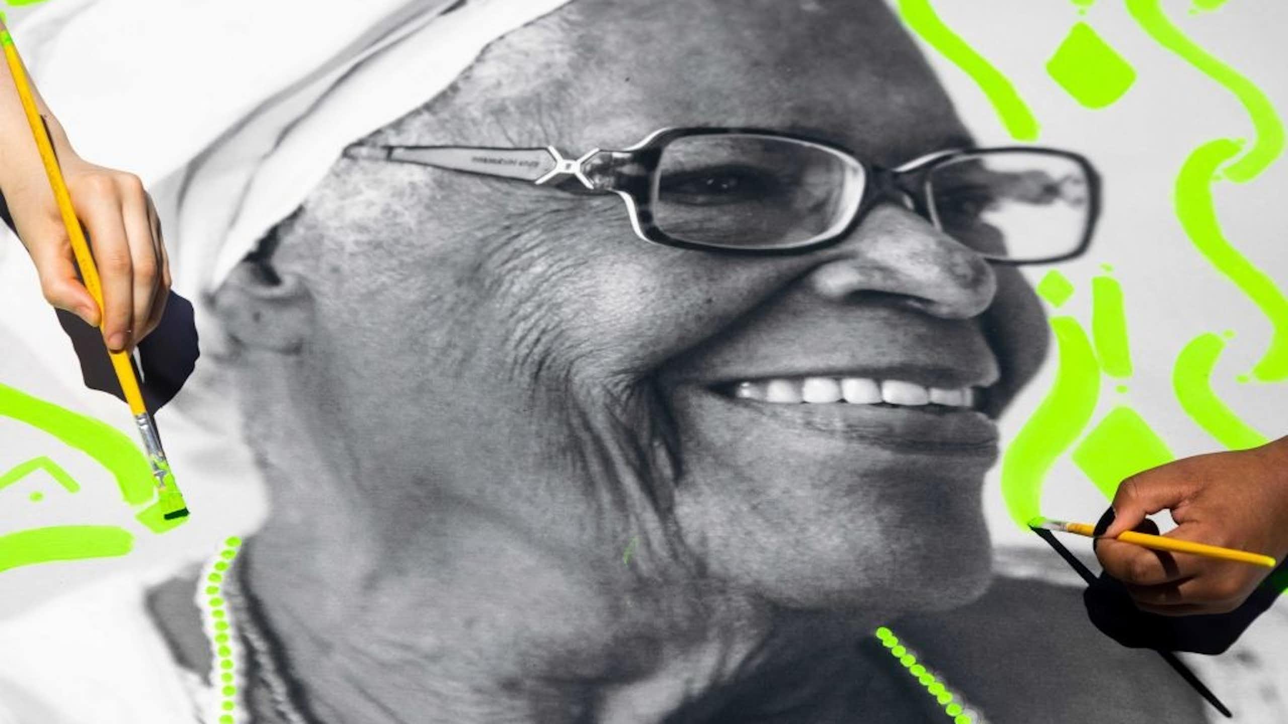 A black and white photo of a smiling elderly woman with hands painting neon green patterns around her.