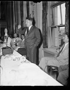 A black and white photo shows a man in a suit, right hand raised, speaking in front of a table.