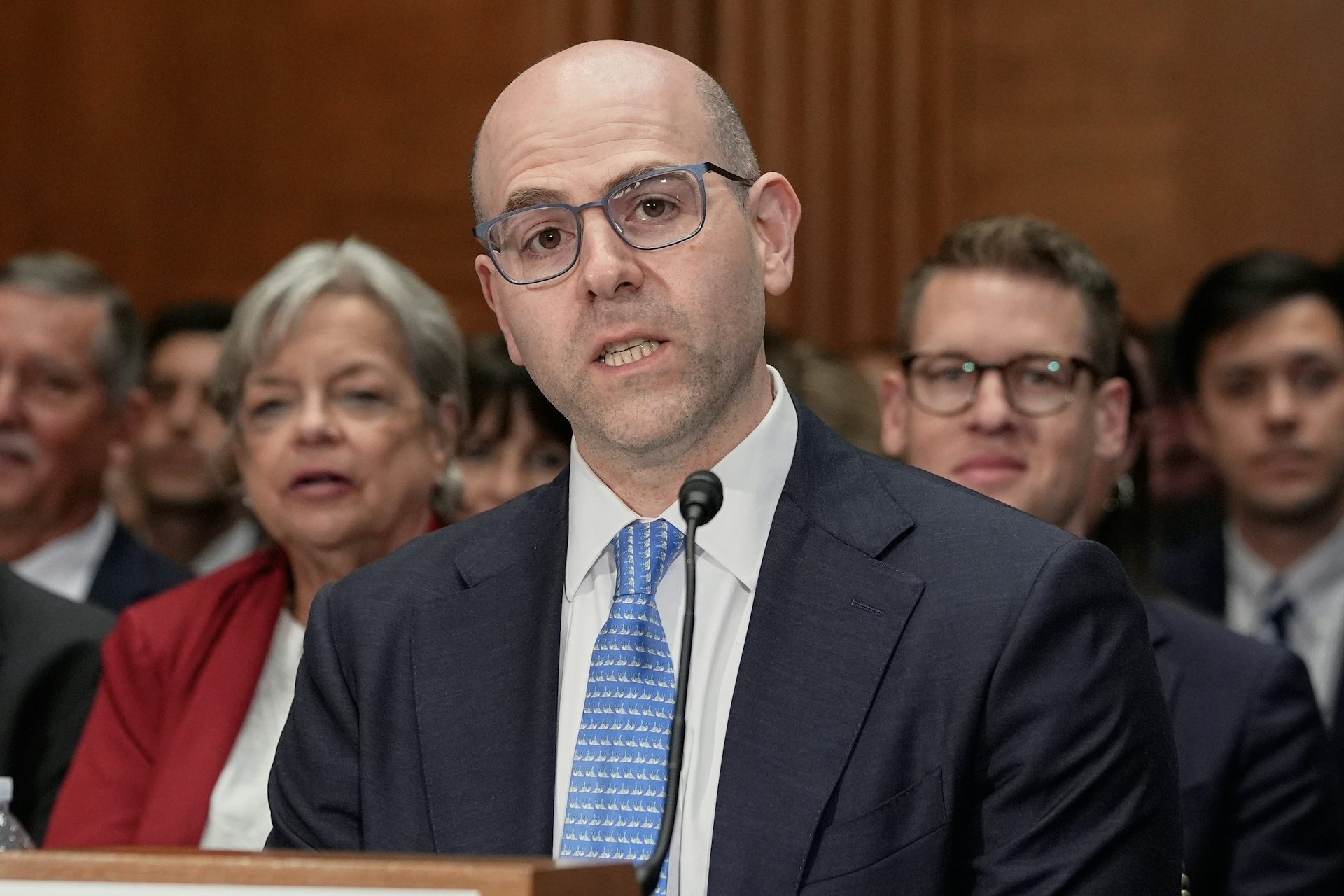 a man in a suit speaks in front of a microphone with a few people sitting in the background