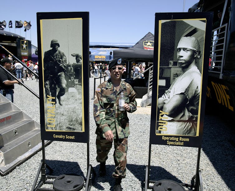 An Army recruiter dressed in military garb stands between two posters depicting Black men in the armed forces.