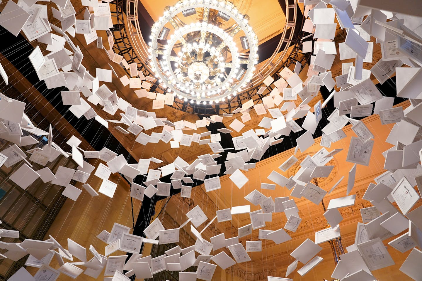 A swirl of white papers hang from a ceiling in an ornate room with a chandelier.