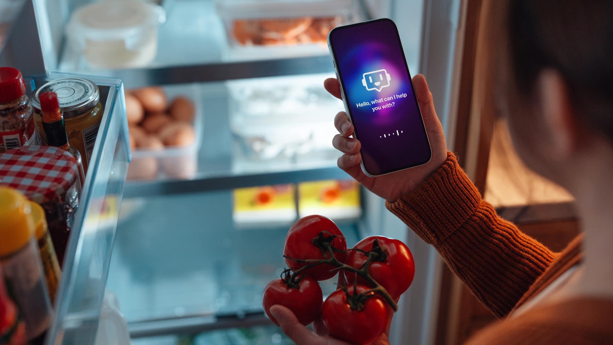 a person in front of an open refrigerator holds a phone in one hand and four tomatoes in the other