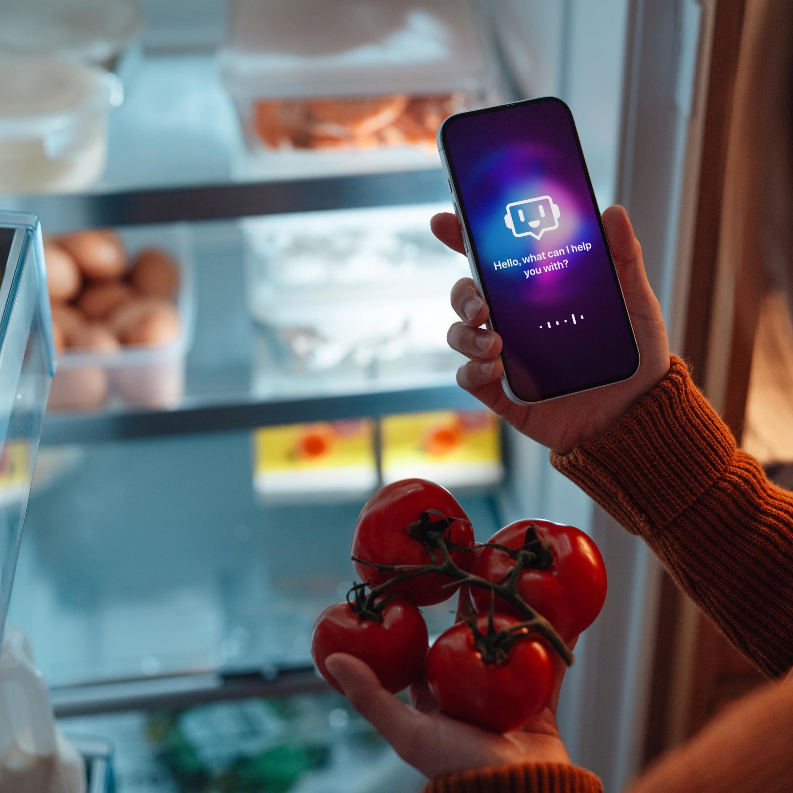 a person in front of an open refrigerator holds a phone in one hand and four tomatoes in the other