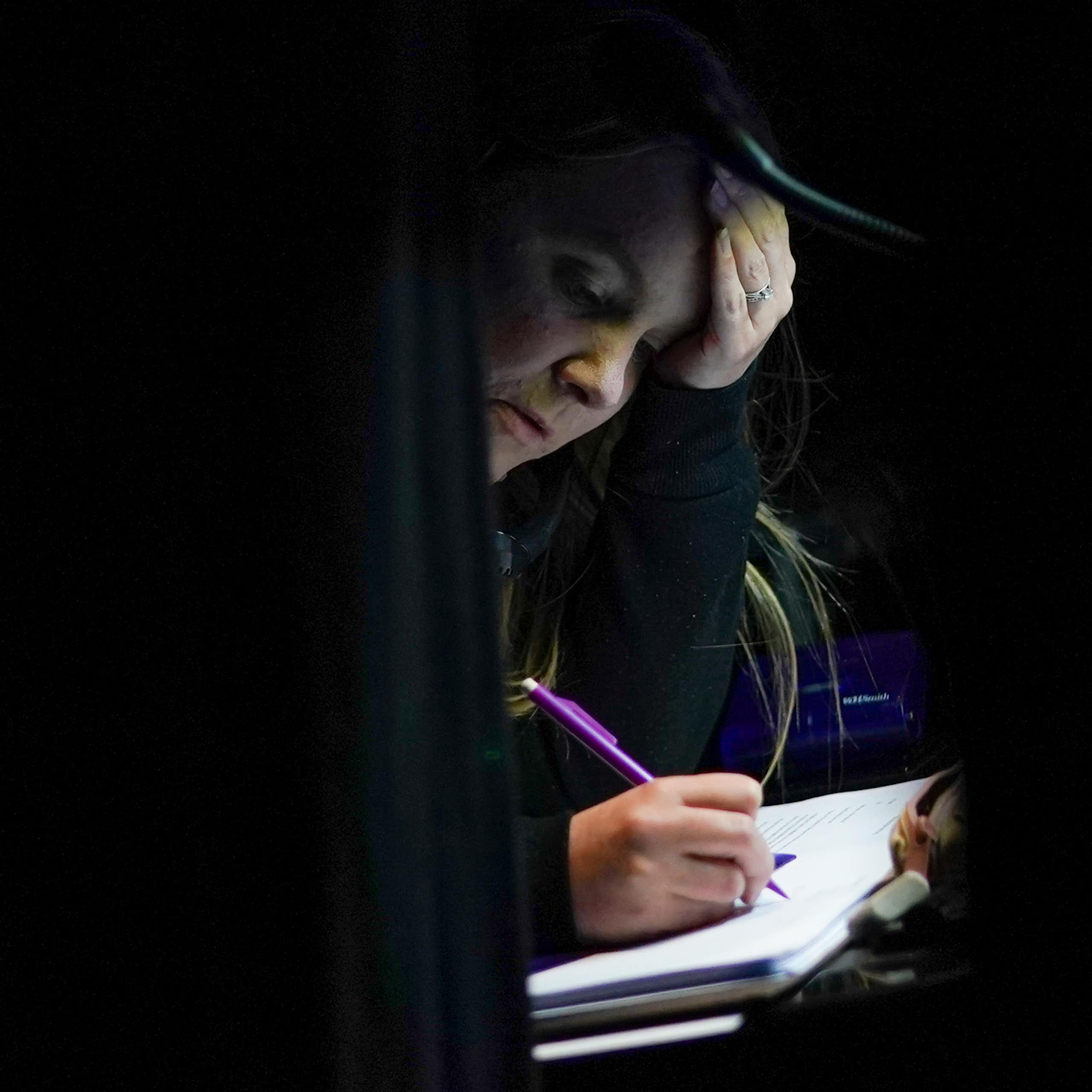 A woman writes in a notebook with a purple pencil in a dark room.
