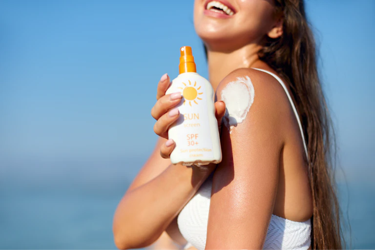 Woman in white bikini applying sun cream on shoulder.