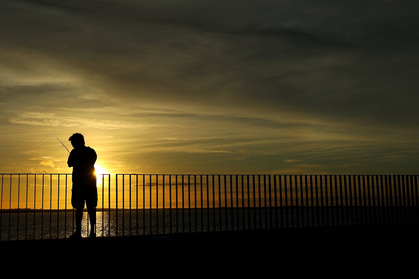 A man stands against a barricade fishing, with the sunrise behind him.