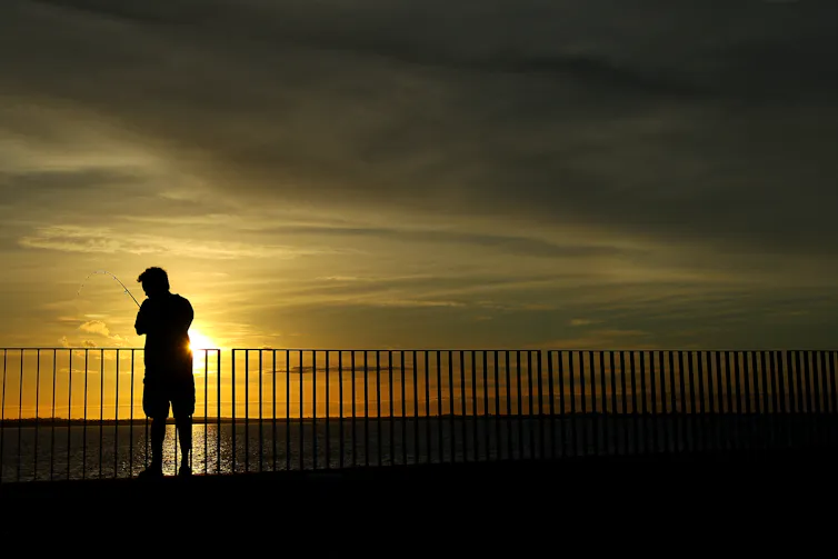 A man stands against a barricade fishing, with the sunrise behind him.