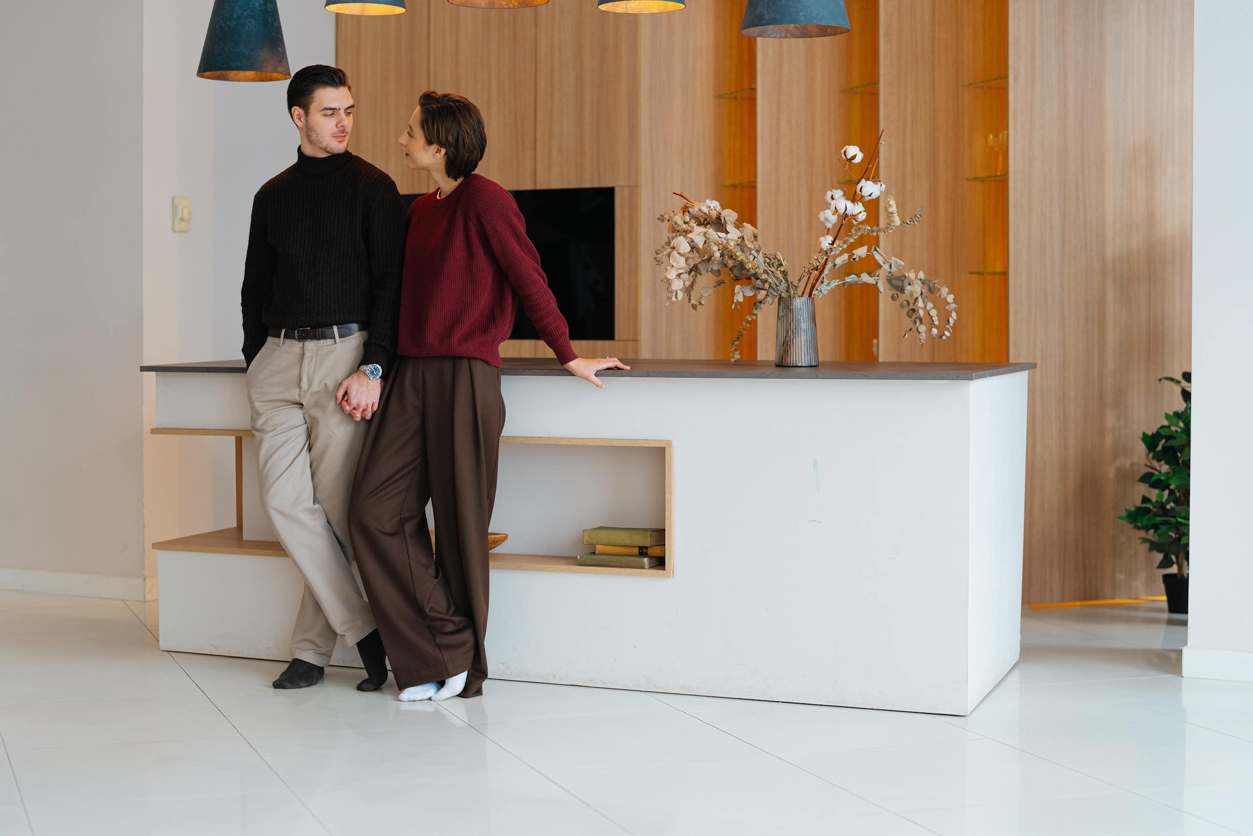 Two adults appear to be looking into each other’s eyes while standing next to each other in an elegant, sparsely furnished room.