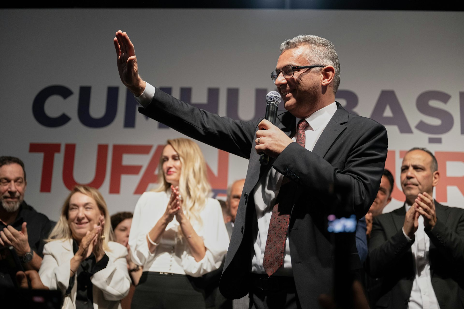 A smiling man wearing glasses with short grey hair holds a microphone and waves.