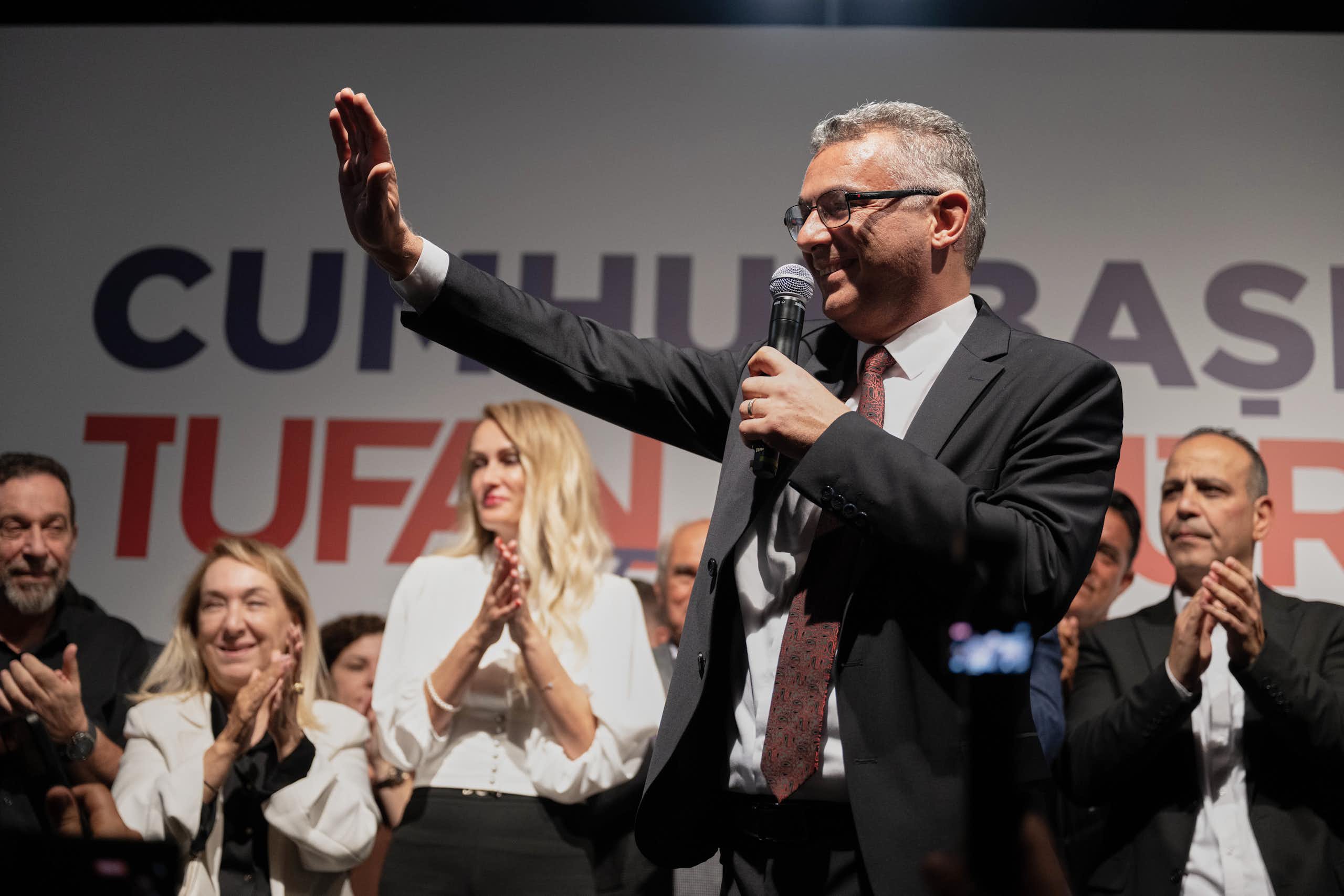 A smiling man wearing glasses with short grey hair holds a microphone and waves.