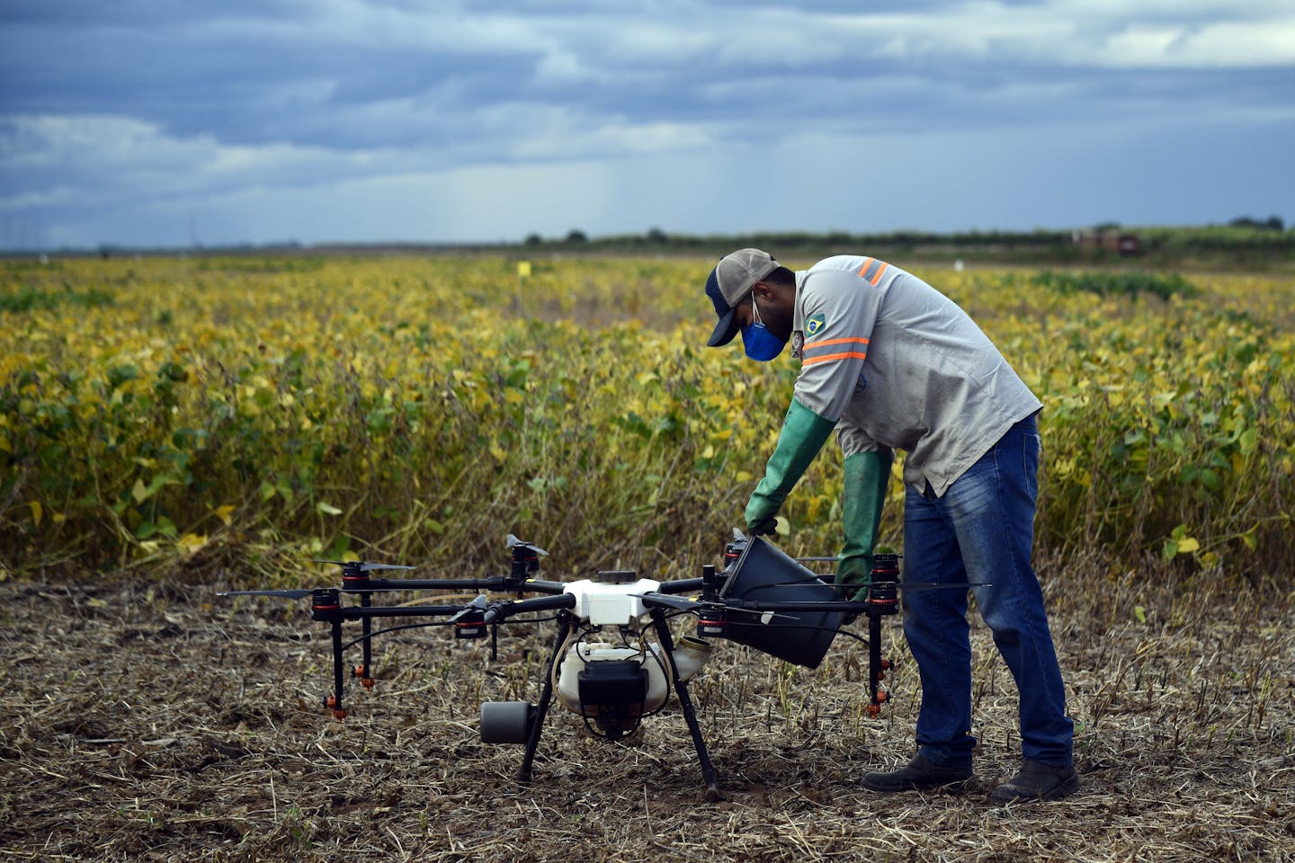 A person pours liquid into a tank attached to a drone, while standing near a large field.