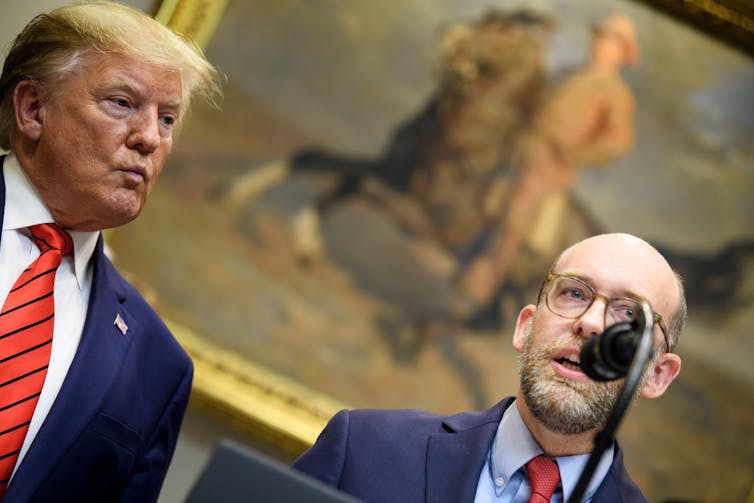 President Trump and Russel Vought stand by a microphone. In the background is a painting of a Theodore Roosevelt on a horse.