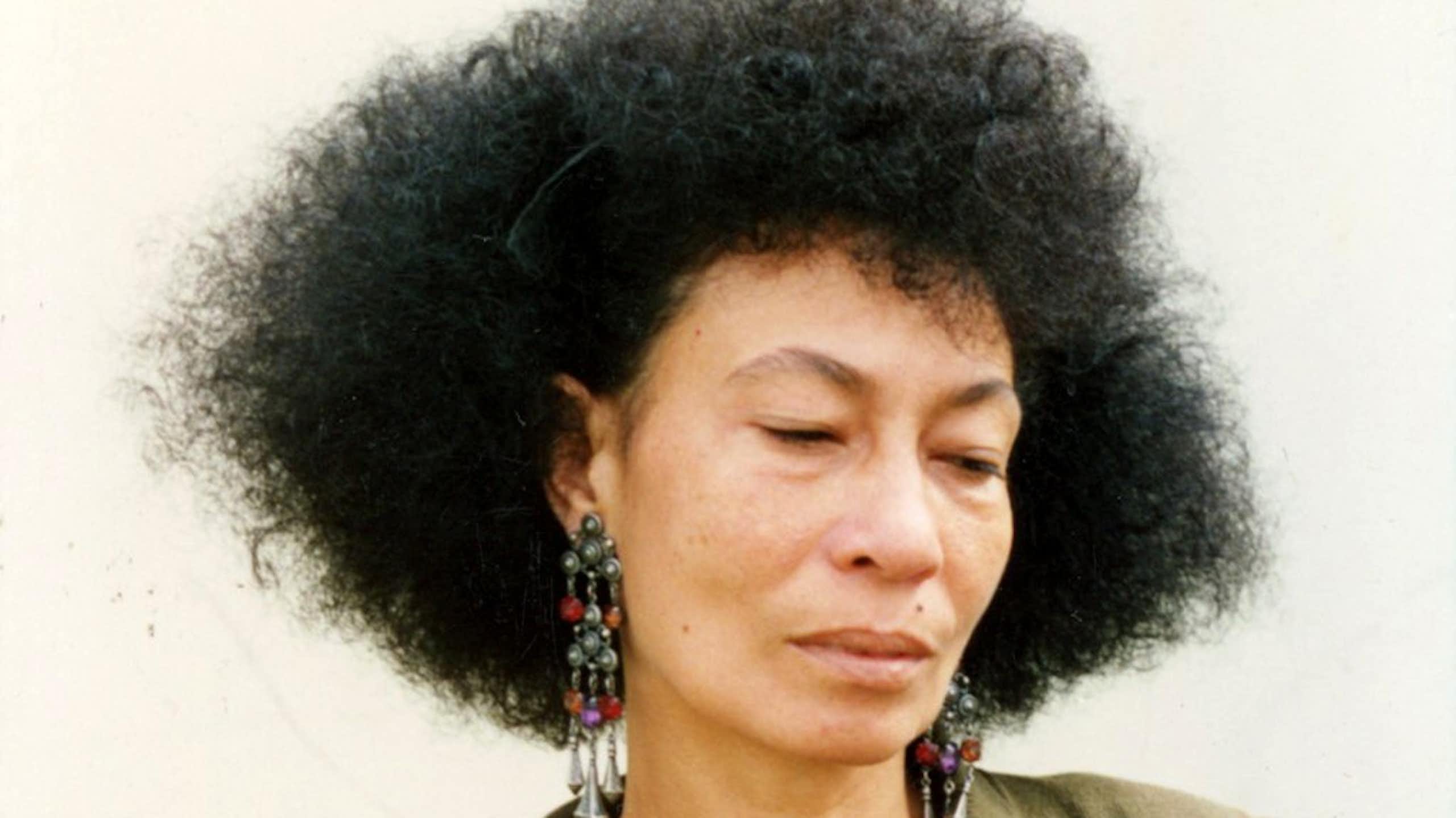 A young light-skinned African woman with a large Afro, eyes cast downward and face deadpan.