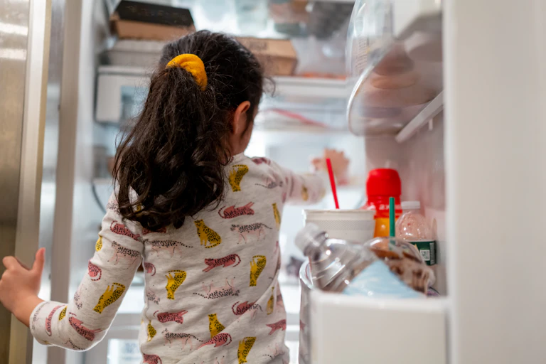 Little girl in pajamas reaches her hand into open refrigerator