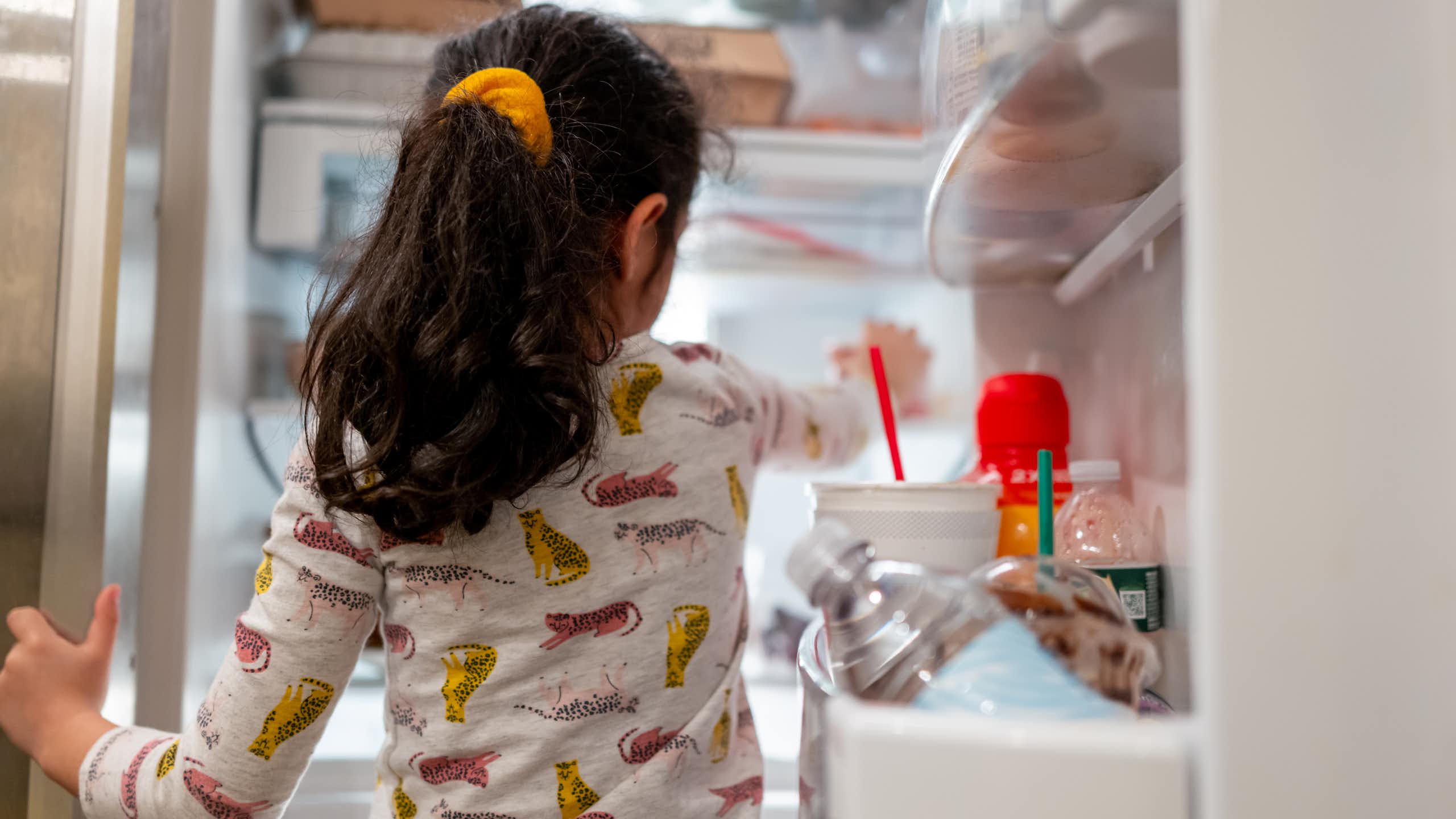 Little girl in pajamas reaches her hand into open refrigerator