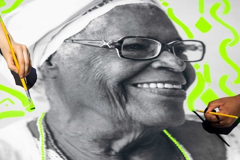 Une photo en noir et blanc d'une femme âgée souriante, les mains peignant des motifs vert fluo autour d'elle.