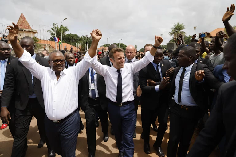 An African man and a European man raise their arms in the air surrounded by a jubilant crowd of African men, mostly in suits.
