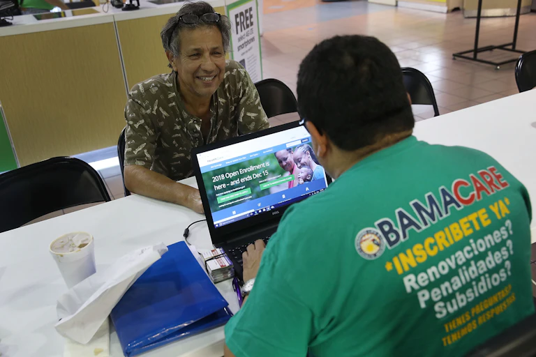 A man sits across from a man with a laptop logged on to healthcare.gov.