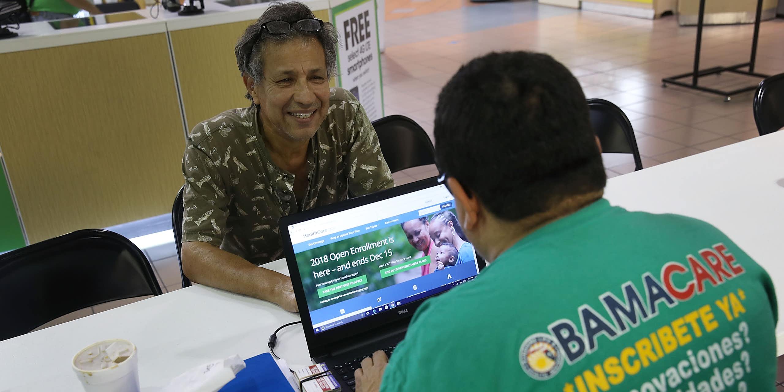A man sits across from a man with a laptop logged on to healthcare.gov.