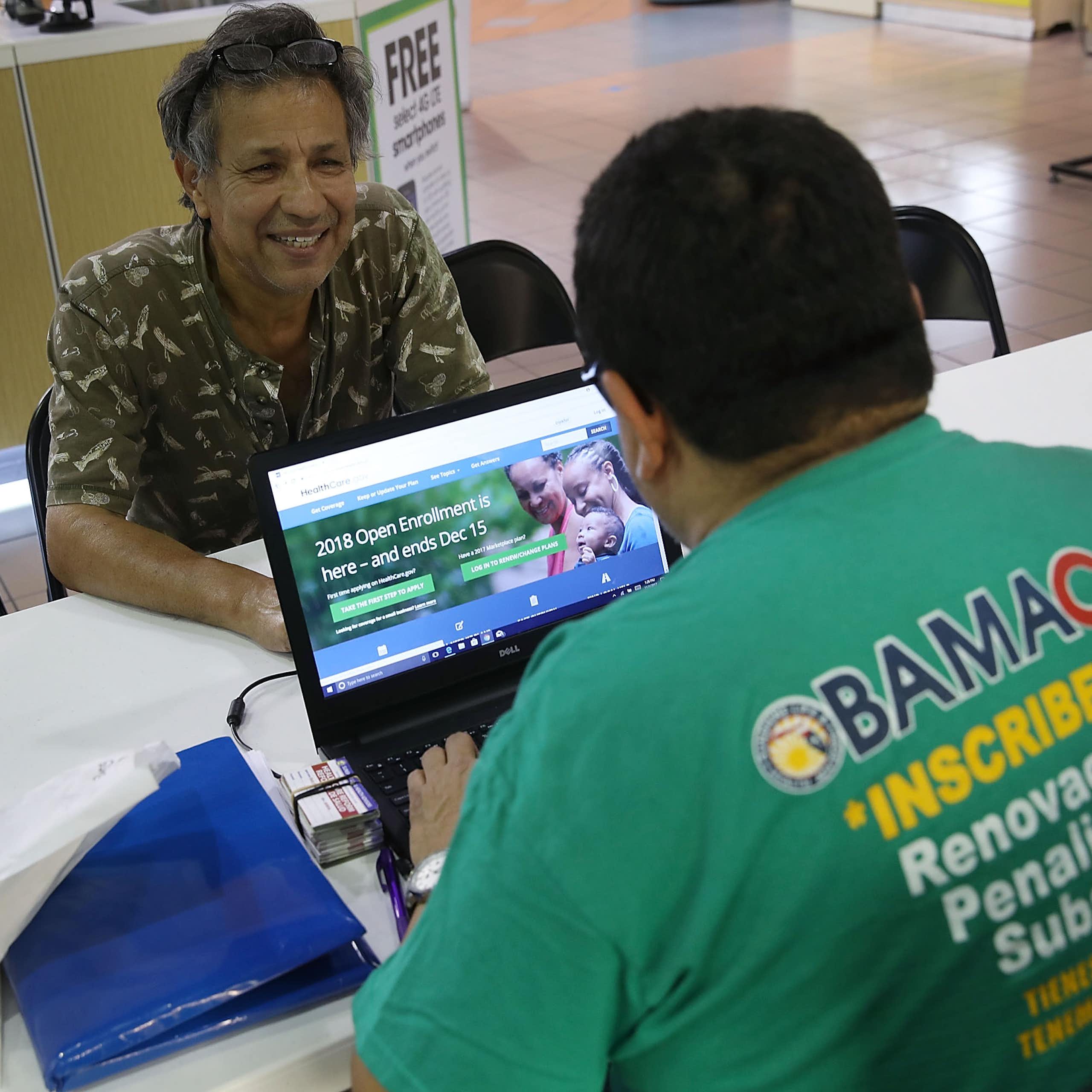 A man sits across from a man with a laptop logged on to healthcare.gov.