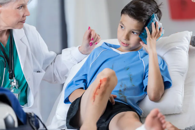 Older female doctor in a lab coat examines a young boy who is holding an ice pack to his head.