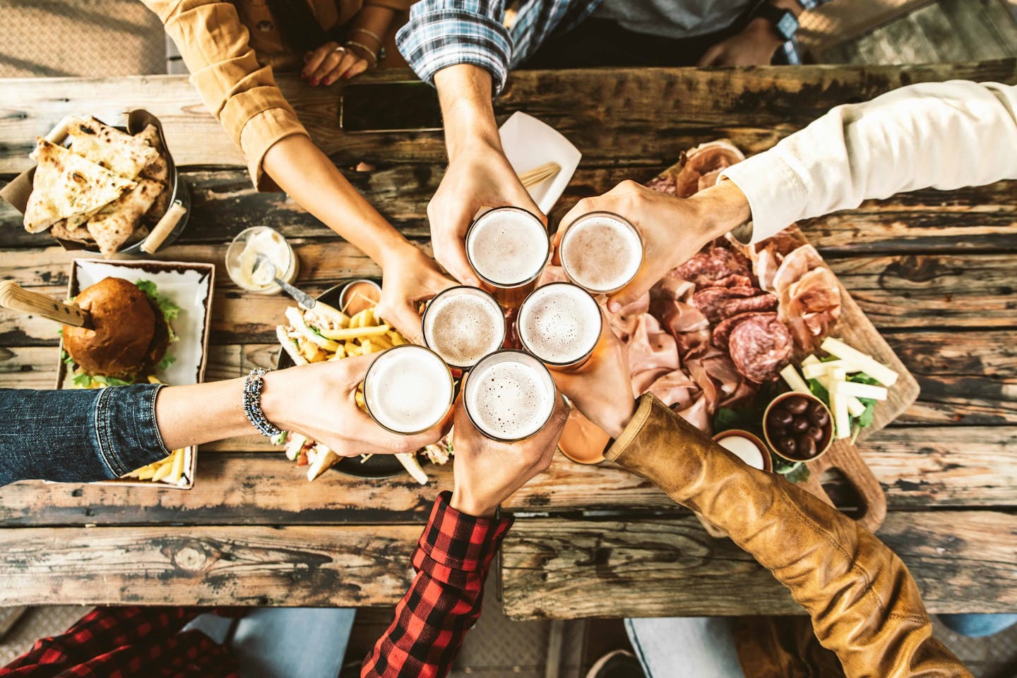 People raising beer glasses at a wooden table covered with food