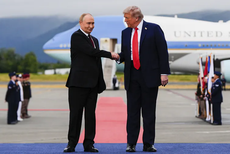 A large man with fluffy blond hair in a blue suit and red tie shakes the hand of a smaller, balding man who is smirking.