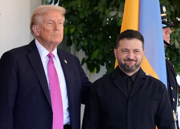 A large man with fluffy orange-ish hair glowers standing next to a shorter man with dark hair and a beard who smiles.