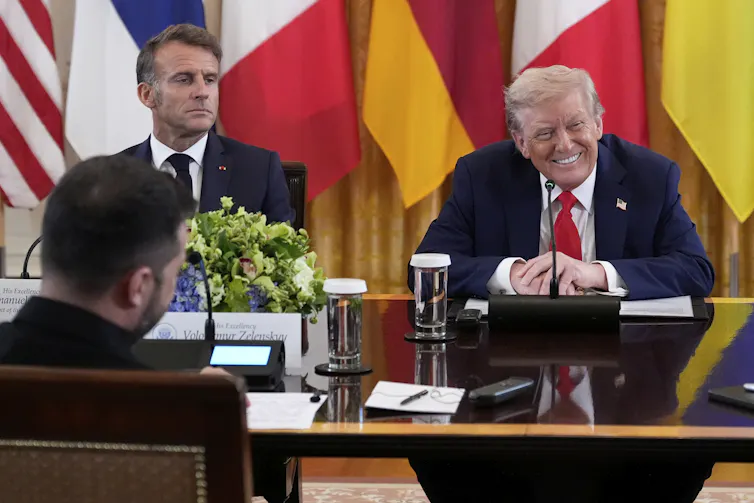 A man with fluffy blond hair smiles as a younger man with dark hair sits next to him looking stern. Another man's back is to the camera.
