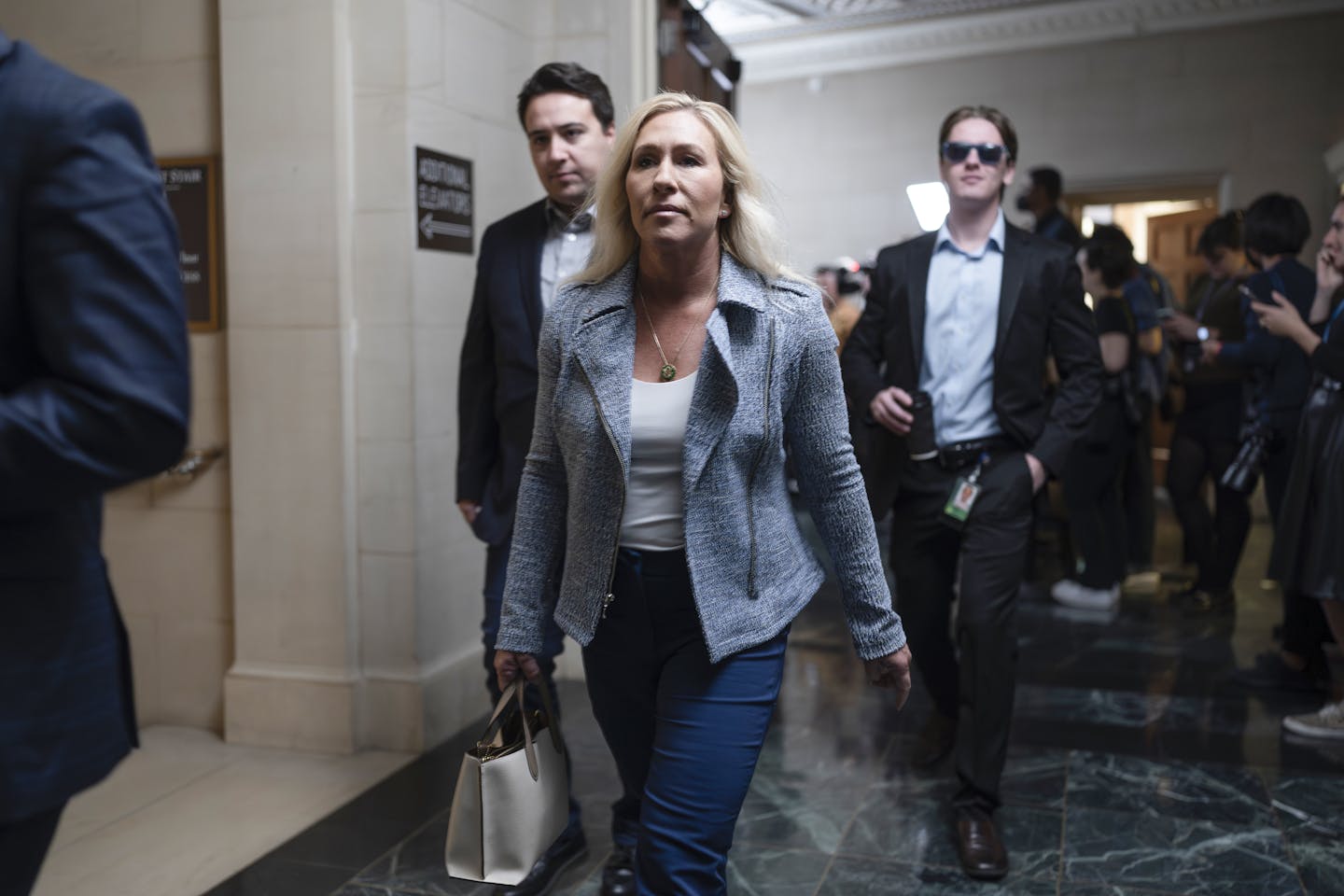 A woman dressed in jeans and a blazer walks down a hall followed by two men.