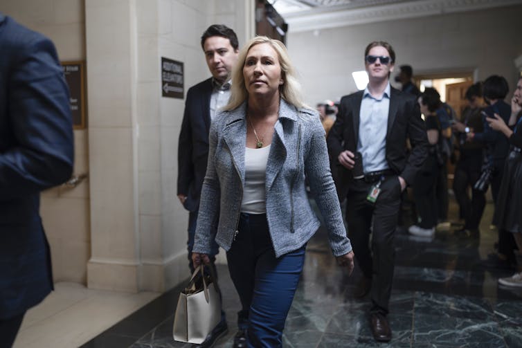 A woman dressed in jeans and a blazer walks down a hall followed by two men.
