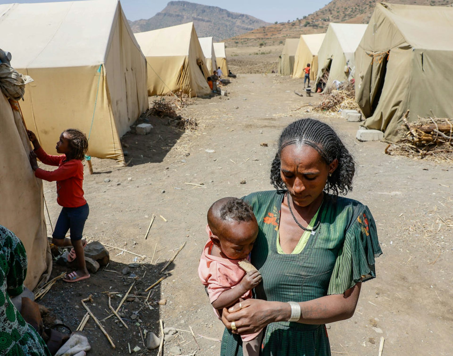 A woman carrying a baby walking on a dusty path between makeshift tents, with children in the background standing outside their tents.
