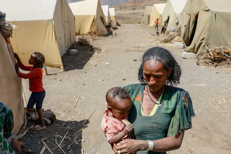 A woman carrying a baby walking on a dusty path between makeshift tents, with children in the background standing outside their tents.