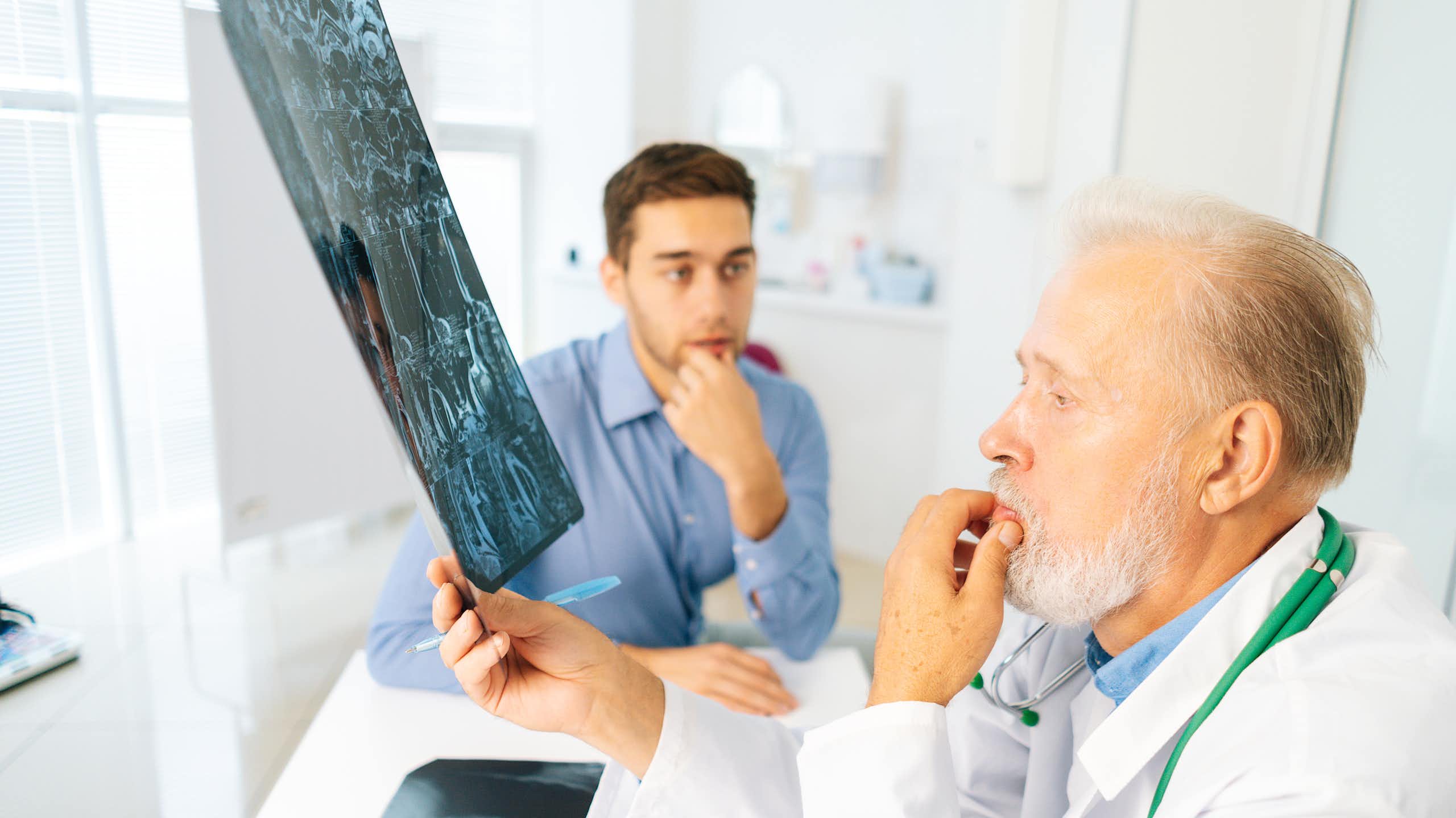 Um médico examina um raio X de um jovem (sentado ao fundo com um olhar preocupado).