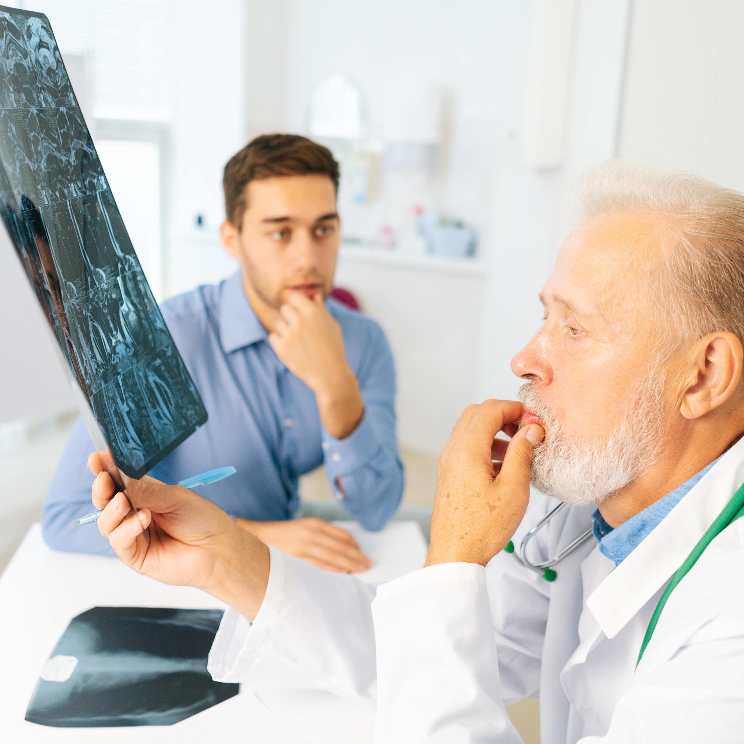 Um médico examina um raio X de um jovem (sentado ao fundo com um olhar preocupado).