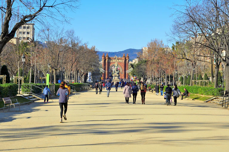 Personas caminando y corriendo por un camino de arena entre los árboles con edificios de una ciudad al fondo