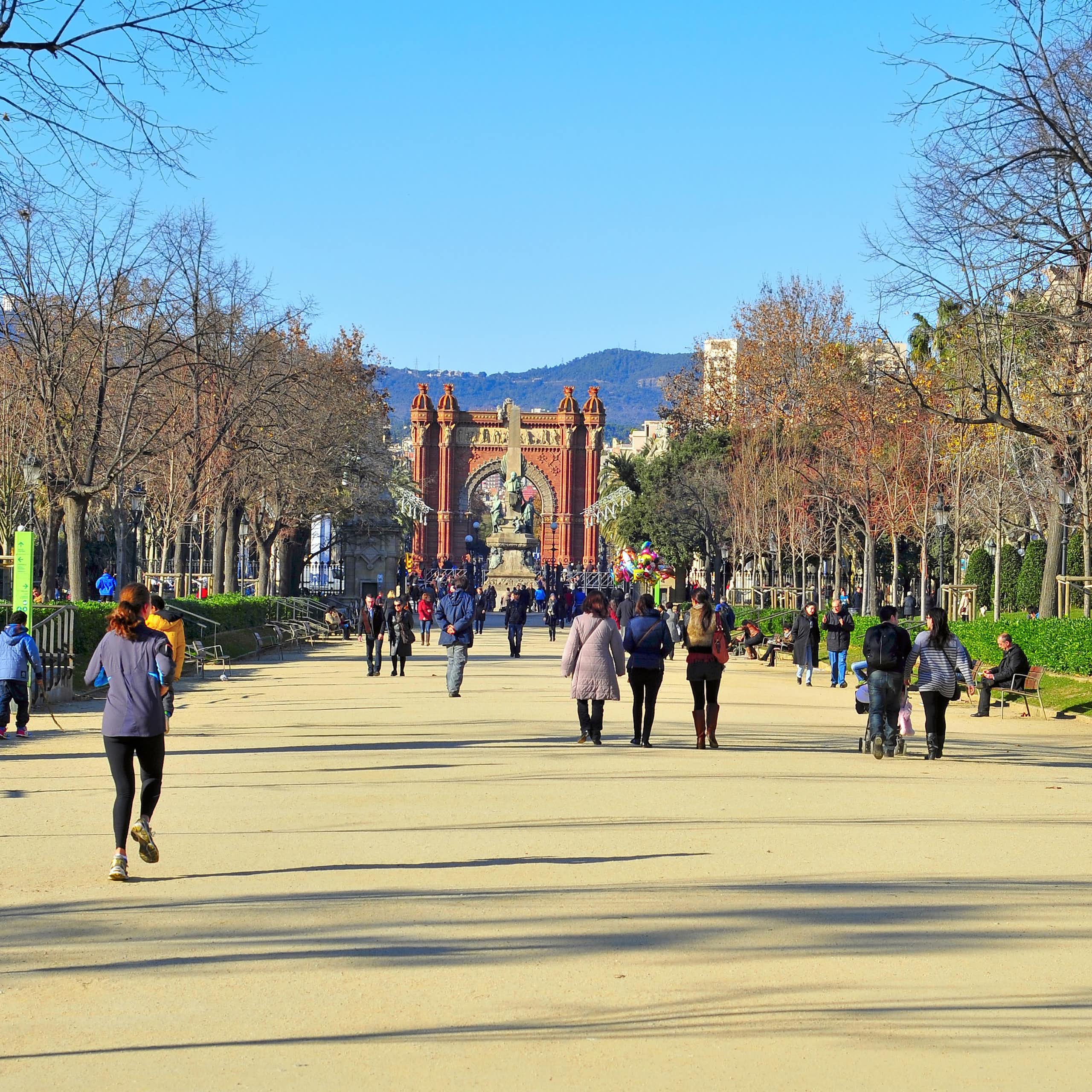 Personas caminando y corriendo por un camino de arena entre los árboles con edificios de una ciudad al fondo
