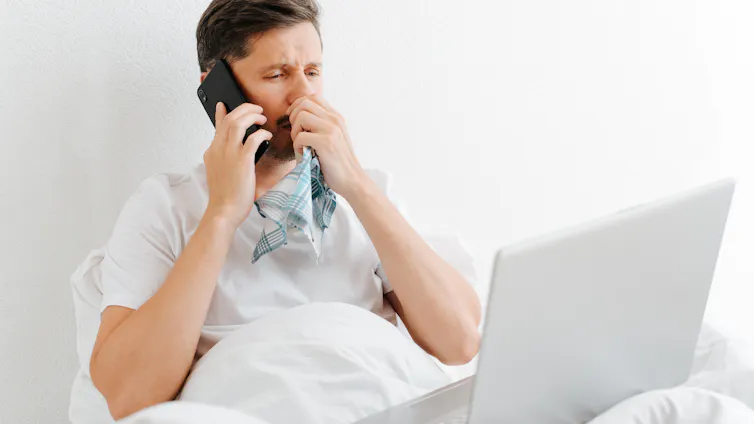 Um homem com sintomas de resfriado na cama olha para um laptop e atende o telefone.